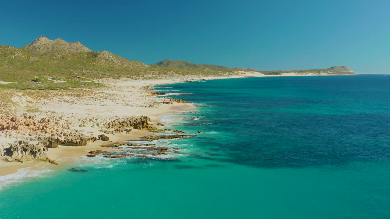 Aerial frame over Buzzards Beach East Cape showing turquoise sea close to shore, a rocky sandy beach curving left to right, distant headland, low hills, and no visible people.