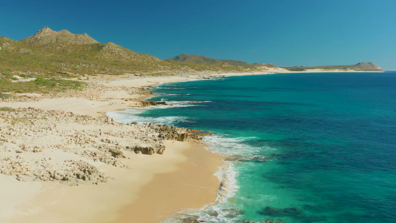 Aerial view over Buzzards Beach in Baja California Sur with pale sand at lower left, turquoise surf curling around rocks, green-brown hills beyond, and no visible people.