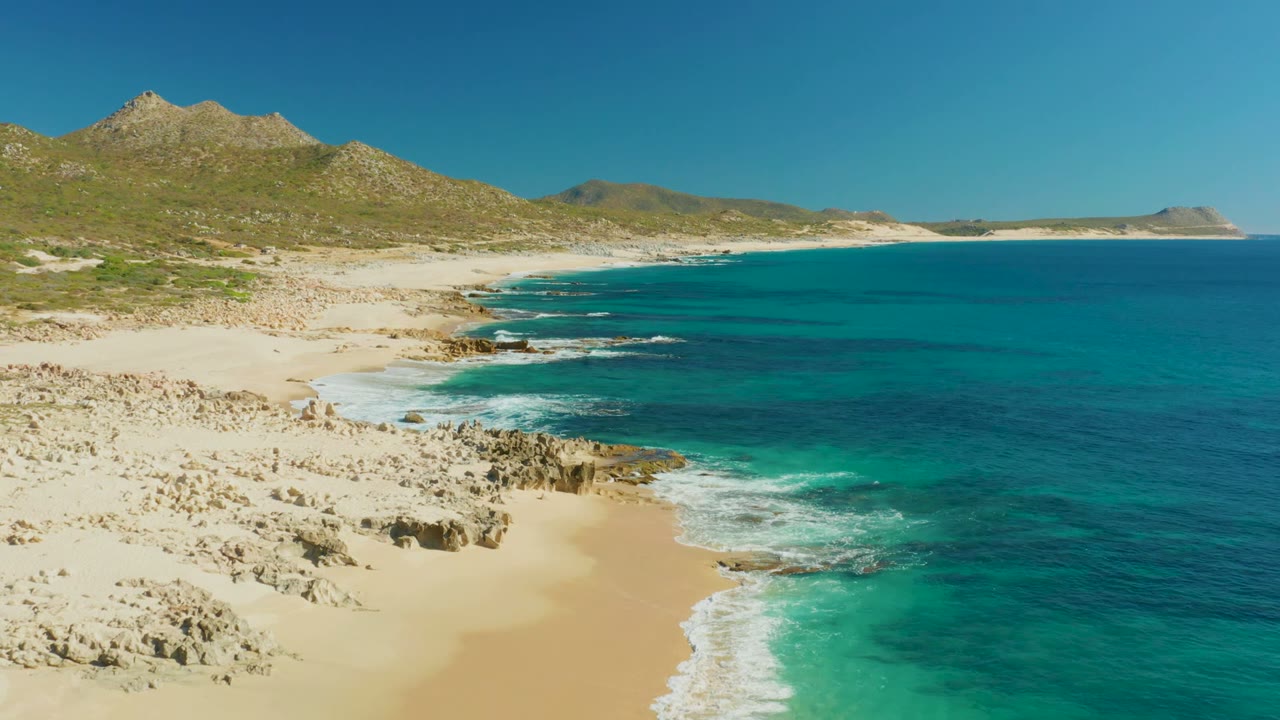 Wide aerial coastline with bright sand, teal water, rocks, and hills.