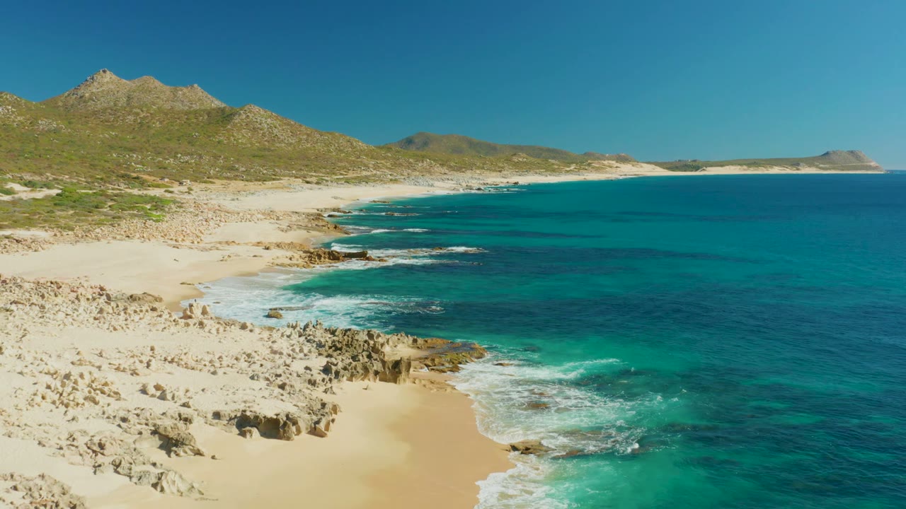 Aerial view at Buzzards Beach with the waterline shifted farther right, white waves breaking near rocky patches, pale beach running toward distant hills, and no visible people.