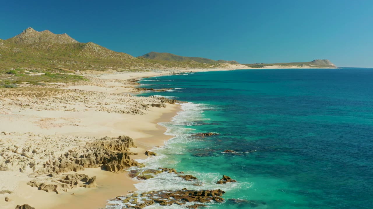 Aerial view over Buzzards Beach East Cape in Baja California Sur, with jagged rocks beside pale sand, turquoise water, low hills, clear sky, and no visible people.