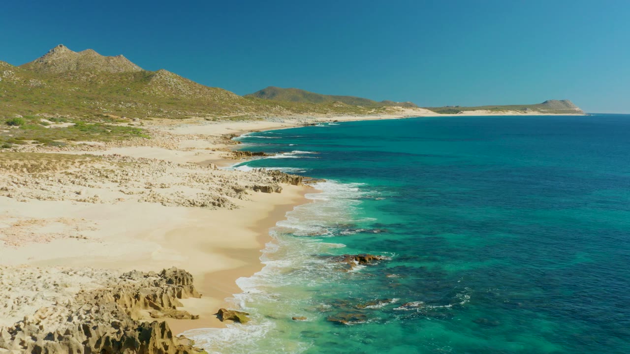 Wide elevated view of Buzzards Beach East Cape, showing blue-green ocean on the right, sandy beach and rock shelves on the left, low hills inland, and no visible people.