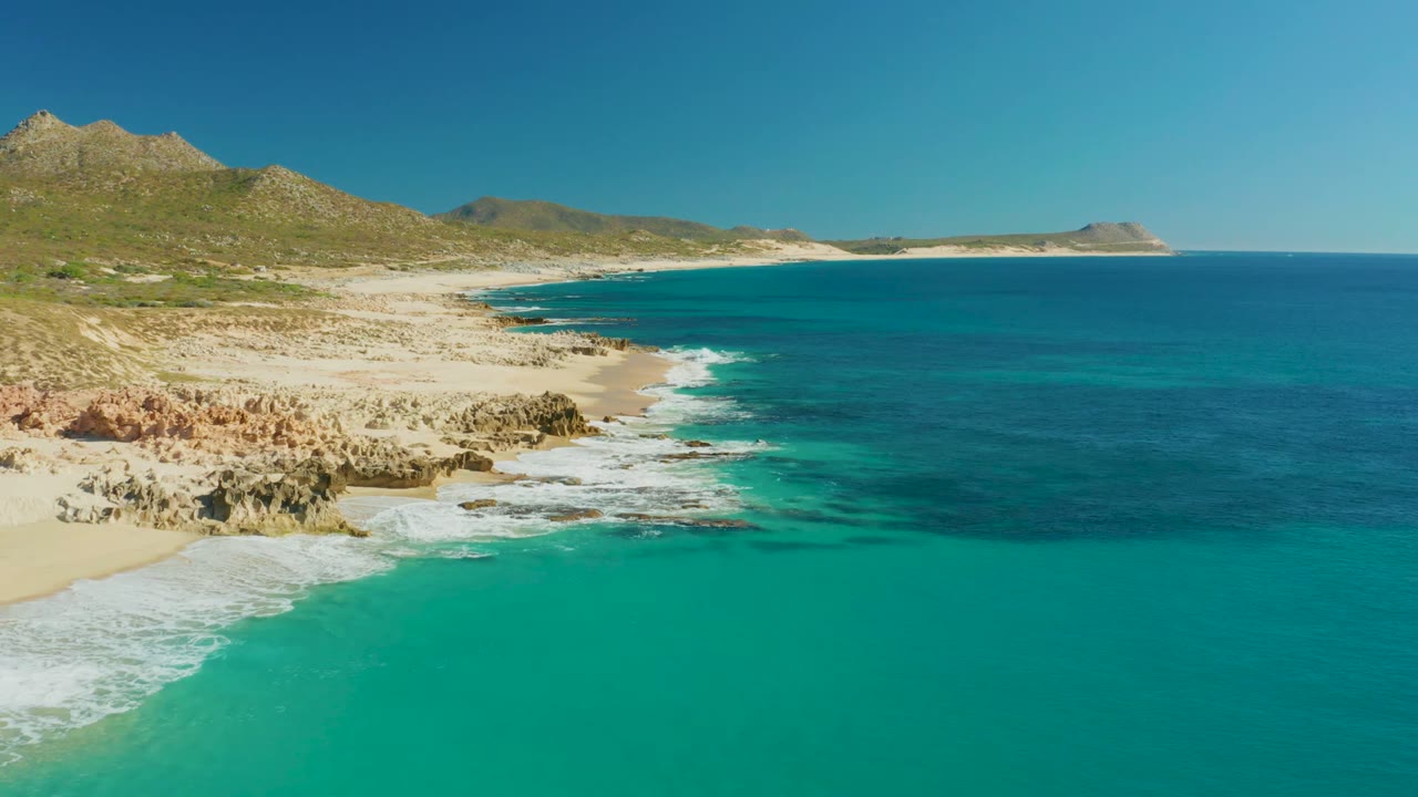 Opening aerial frame over Buzzards Beach in Baja California Sur, with turquoise surf washing onto pale sand and rocks, low hills behind the coast, and no visible people.