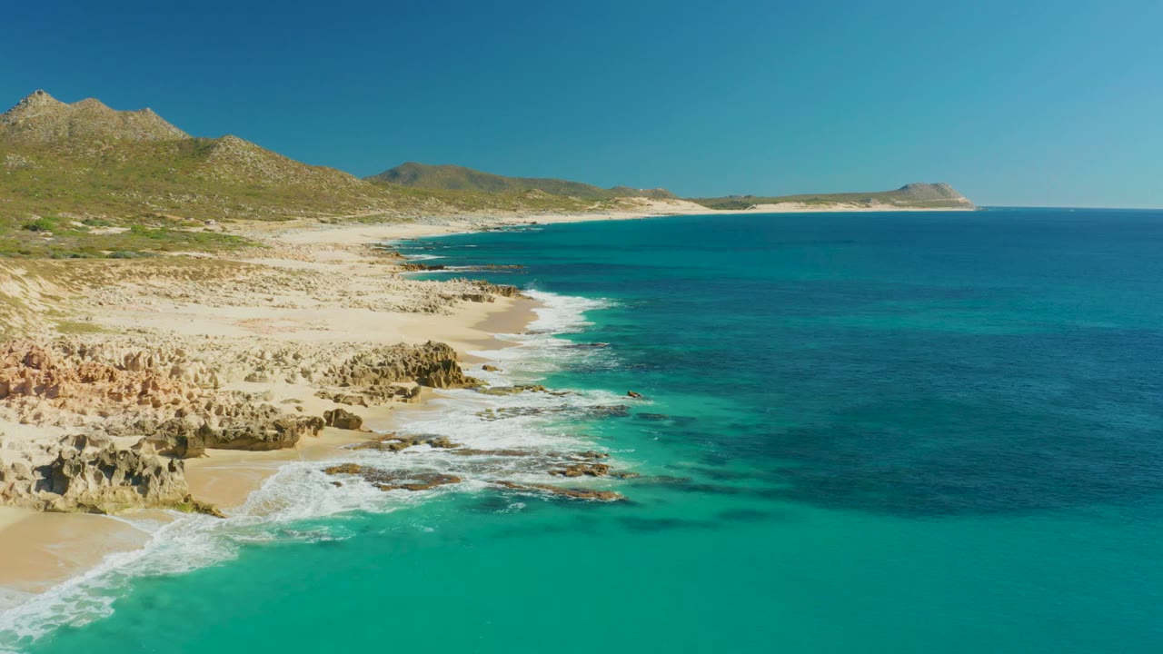 Buzzards Beach coastline seen from above, with foamy water crossing the rocky shore, turquoise shallows in front, darker ocean to the right, hills in back, and no visible people.