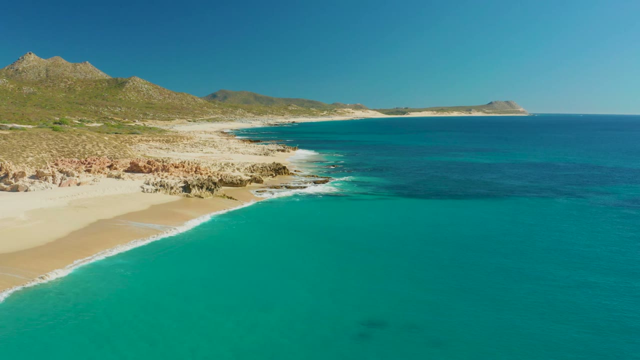 Opening frame over Buzzards Beach East Cape in Baja California Sur, with turquoise water in the foreground, a sandy cove at left, rocky outcrops along the shore, dry hills behind, and no visible people.