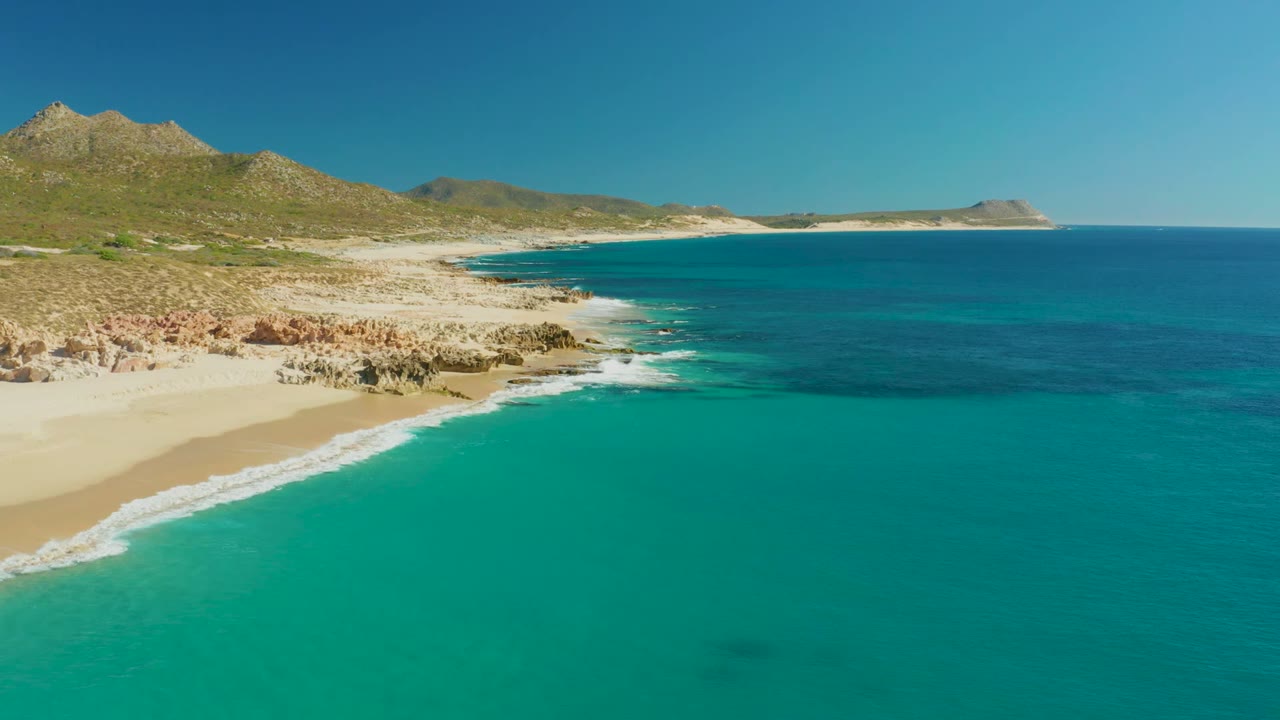 Turquoise ocean and white surf along a sandy beach with hills beyond