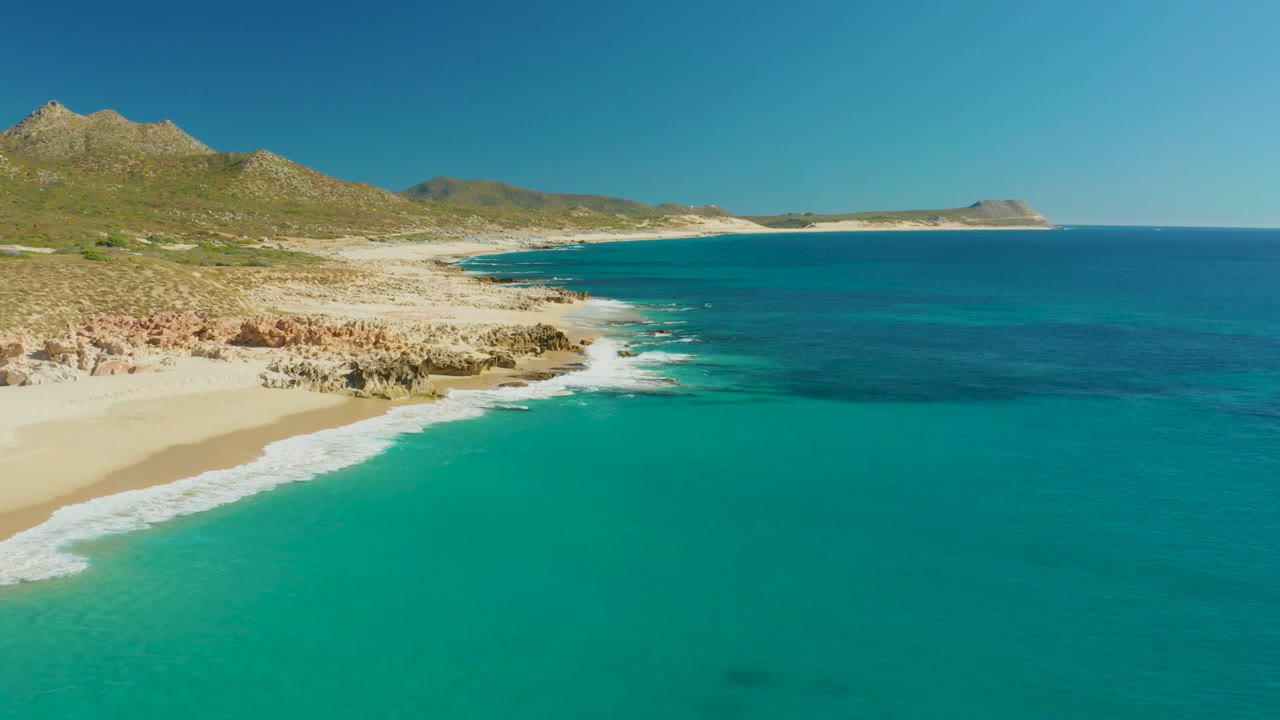 Aerial coastal frame from Buzzards Beach East Cape with the sandy beach left of frame, rocky outcrops tracing the waterline, a broad turquoise foreground, dry hills inland, and no visible people.