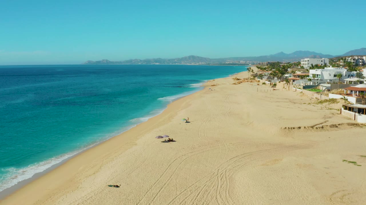 Aerial view over Buzzards Beach East Cape BCS Mexico with turquoise water on the left, a broad sandy shoreline, small beach umbrellas, distant people, and beachfront buildings to the right.