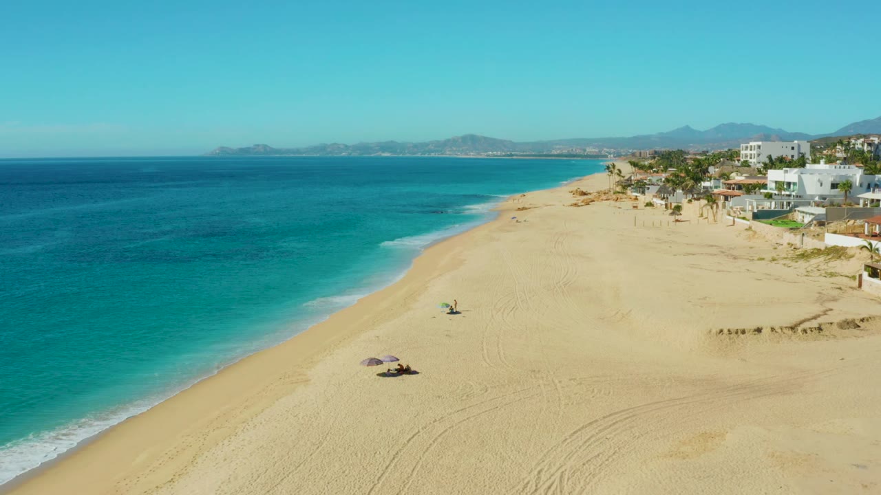 Wide aerial shoreline with blue ocean, pale sand, small umbrellas, and buildings along the beach.