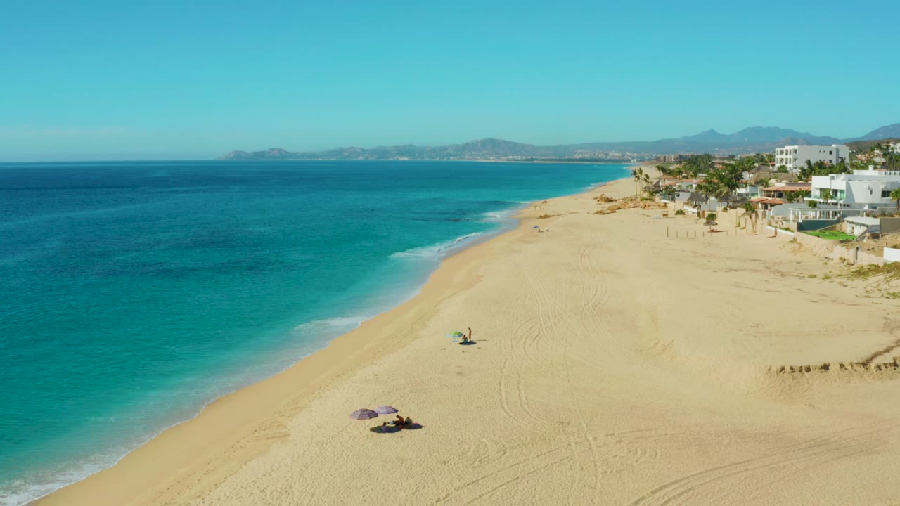 High aerial frame of Buzzards Beach with the wave line farther up the left shore, two umbrella areas on the sand, small people nearby, and beachfront buildings along the upper right.