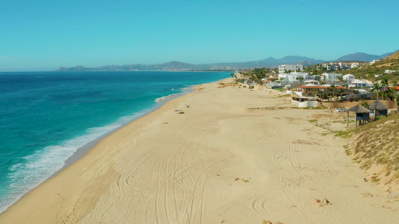 Wide aerial view of Buzzards Beach in Baja California Sur with turquoise ocean on the left, broad sand in front, beachfront buildings on the right, distant mountains, and small people visible on the beach.