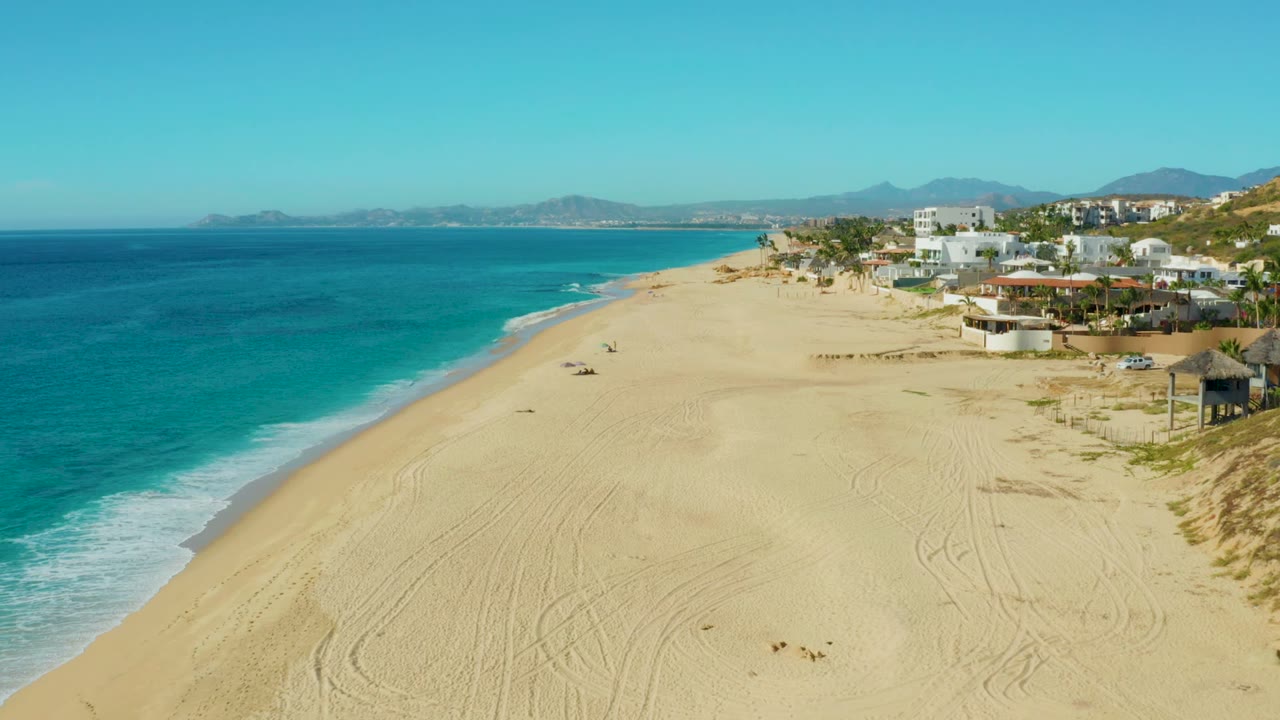 Elevated coastline view with turquoise water, pale sand, distant hills, and buildings along the beach.