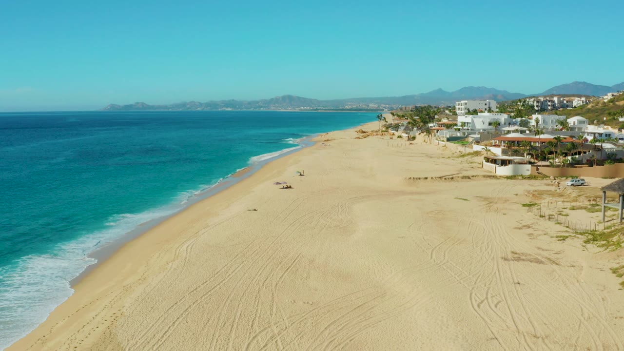 Aerial frame over Buzzards Beach showing white surf curling along the left edge, open raked sand below, small people near the center, and homes climbing the right side toward low mountains.