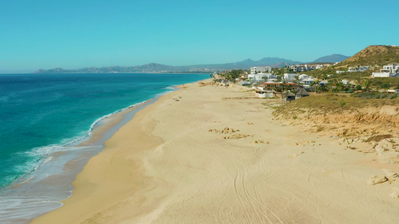 Opening aerial view at Buzzards Beach East Cape, Baja California Sur, with turquoise water on the left, a broad empty sandy beach, dunes, coastal buildings, distant hills, and no visible people.