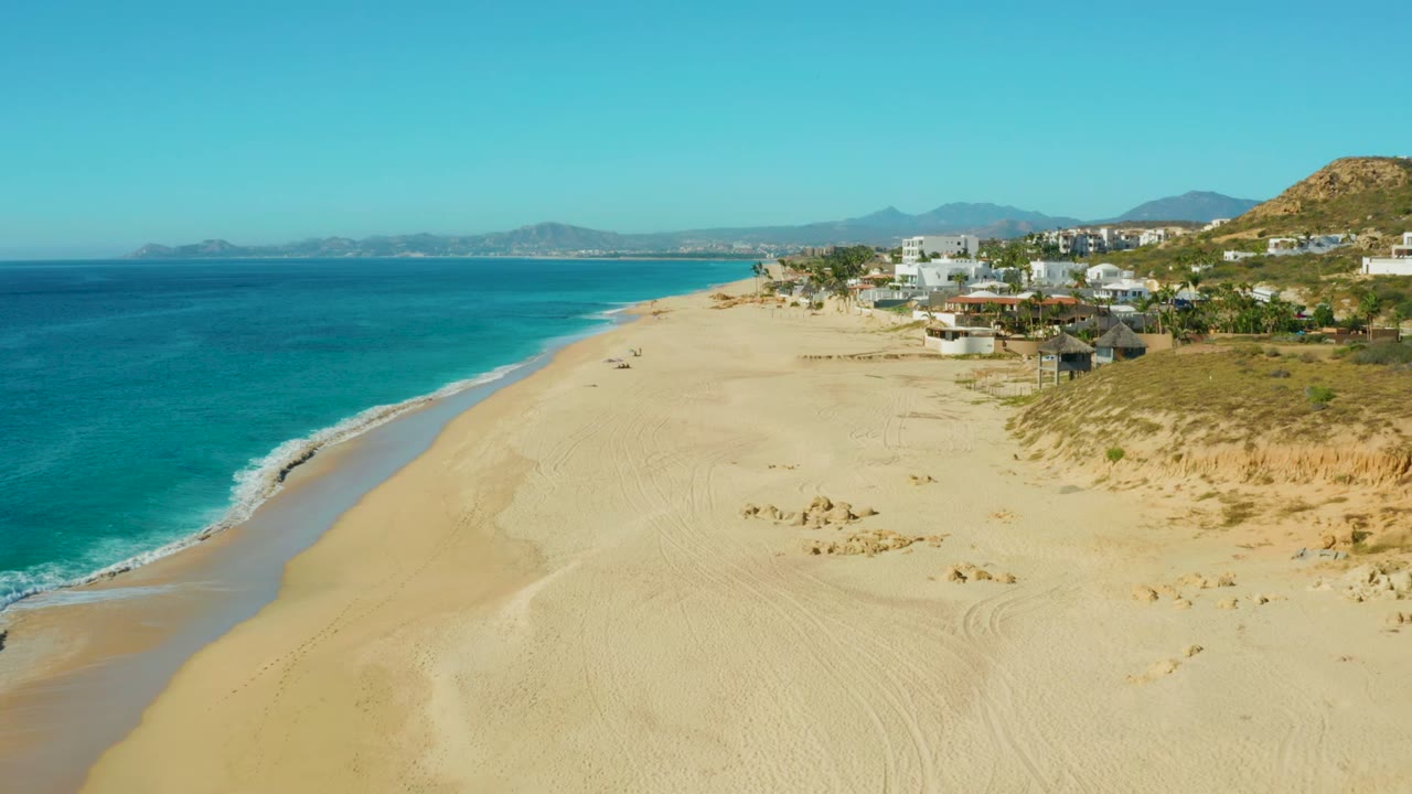 At 0:02, the aerial view holds the Buzzards Beach East Cape shoreline with turquoise water left, smooth sand center, dunes and clustered coastal buildings right, low hills behind, and no visible people.