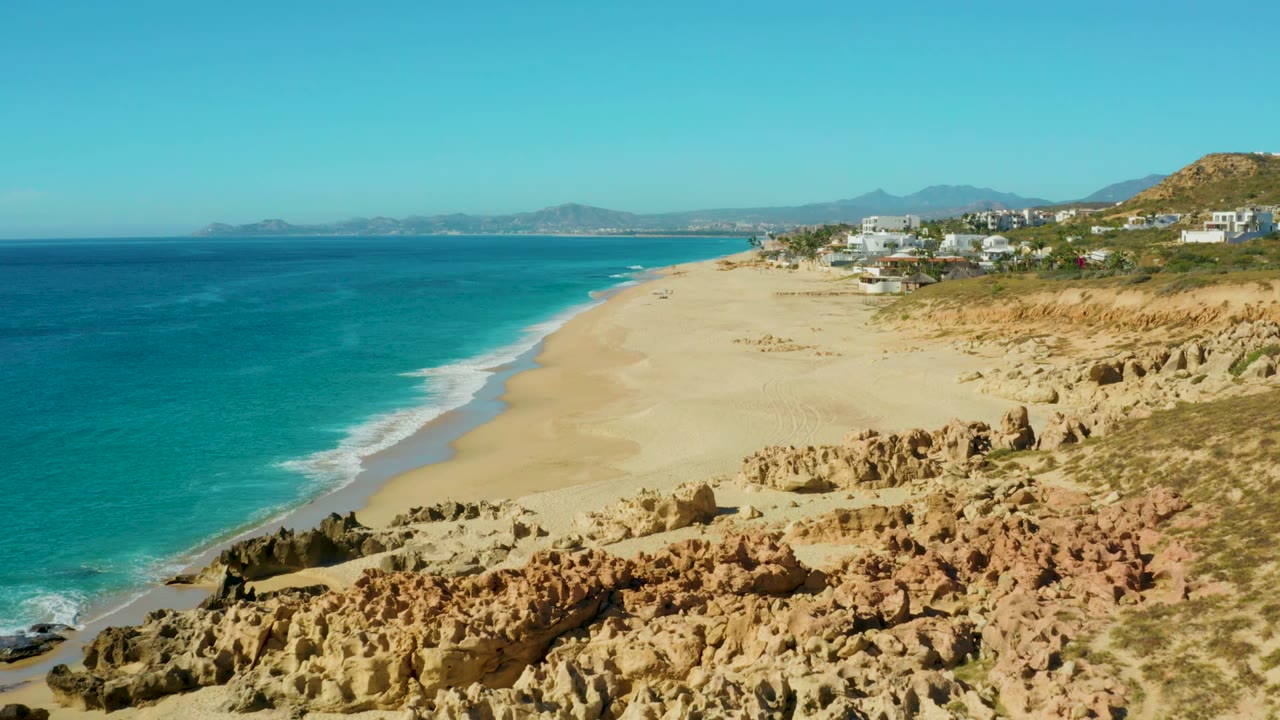 Aerial view at Buzzards Beach in Baja California Sur with turquoise water on the left, pale sand through the center, tan rocks in the foreground, distant buildings, and no visible people.