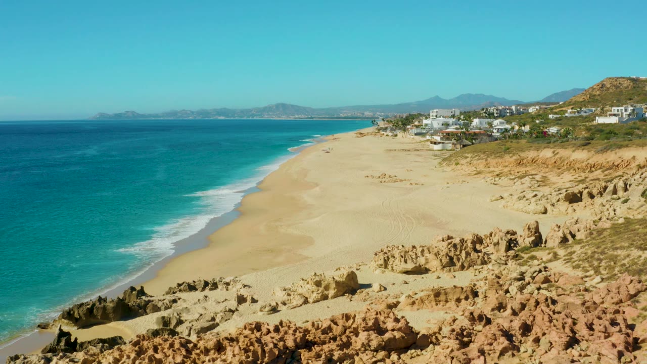 Wide beach aerial with surf line, tan rocks, and coastal buildings.