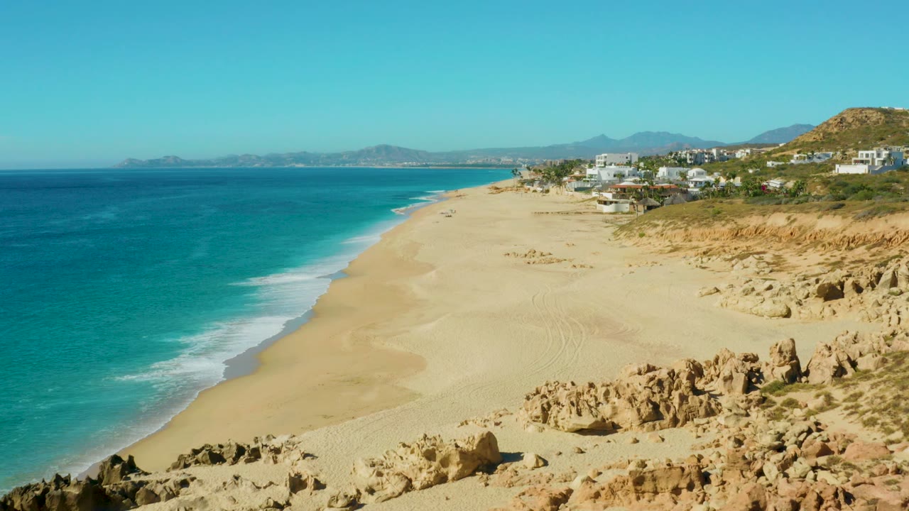 Aerial view of Buzzards Beach with deeper blue water at far left, bright turquoise shallows, a broad empty sand area, eroded cliffs at right, and no visible people.