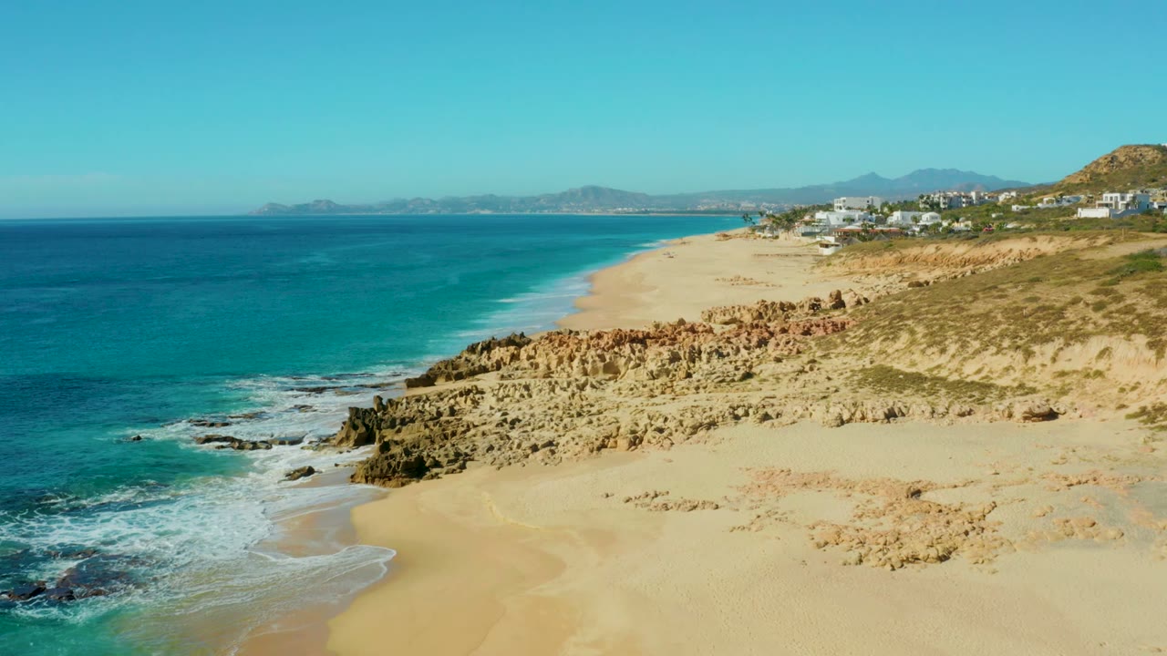 Opening aerial frame over Buzzards Beach East Cape Los Cabos, with turquoise water left, pale sand curving inland, rocky cliffs in front, distant hillside buildings, and no visible people.
