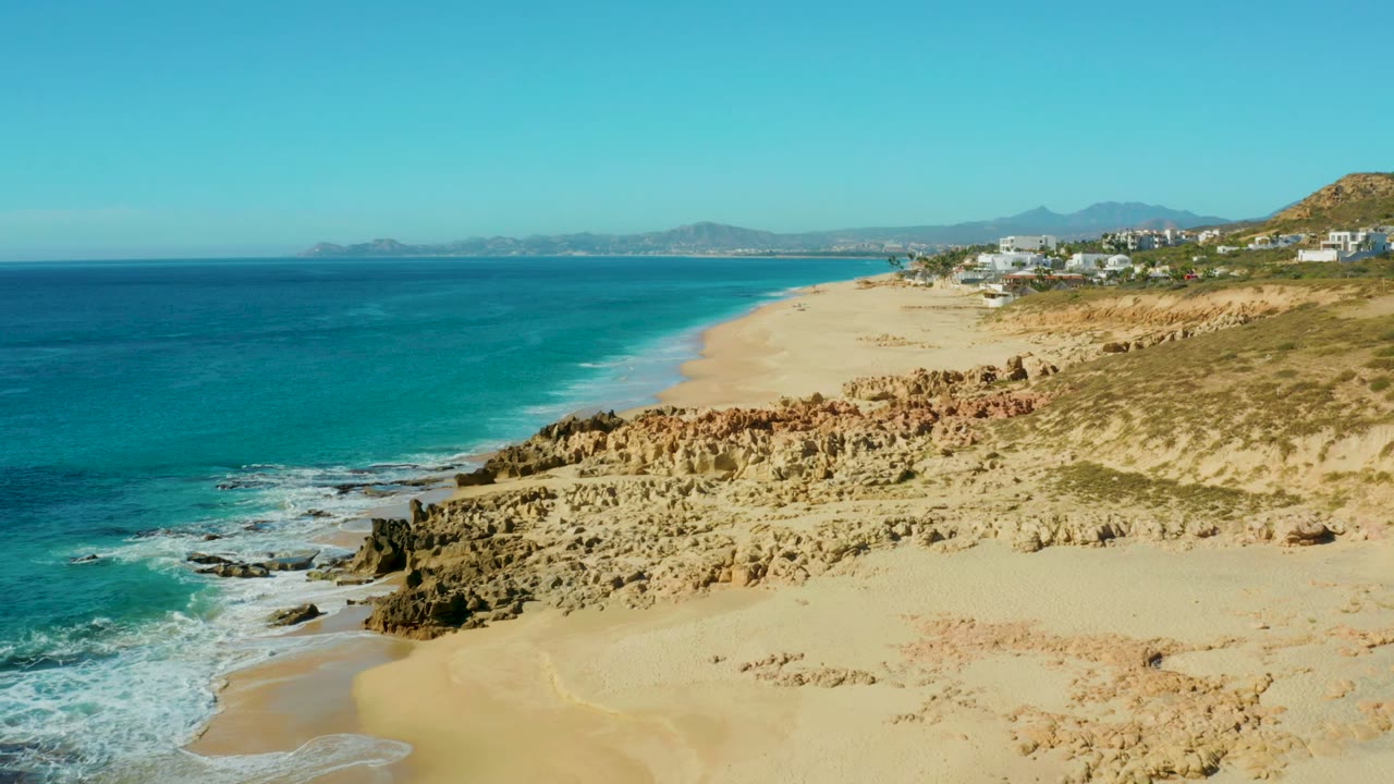 Buzzards Beach shoreline from above at 0:01, showing foamy waves against wet sand, tan cliff formations across the lower frame, distant hills, small buildings, and no visible people.
