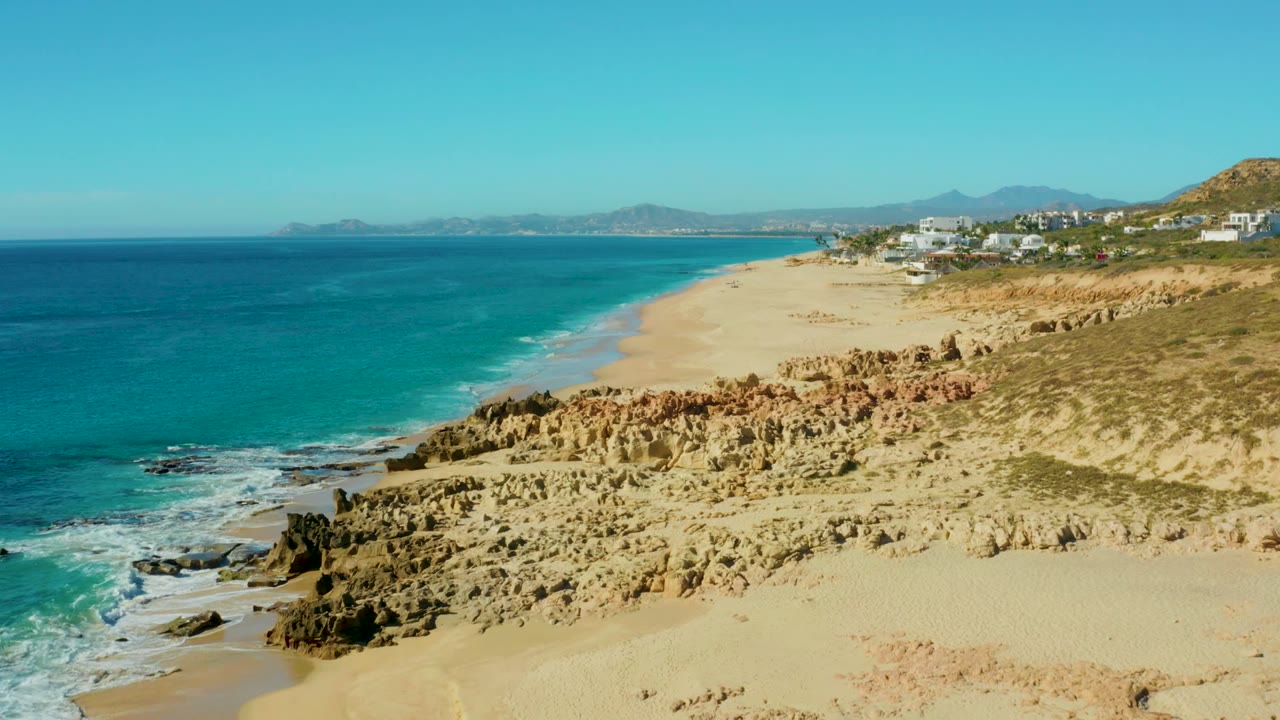 Aerial frame of Buzzards Beach with blue-green water on the left, a broad empty beach running toward distant hills, rough tan rock shelves in the foreground, hillside buildings, and no visible people.