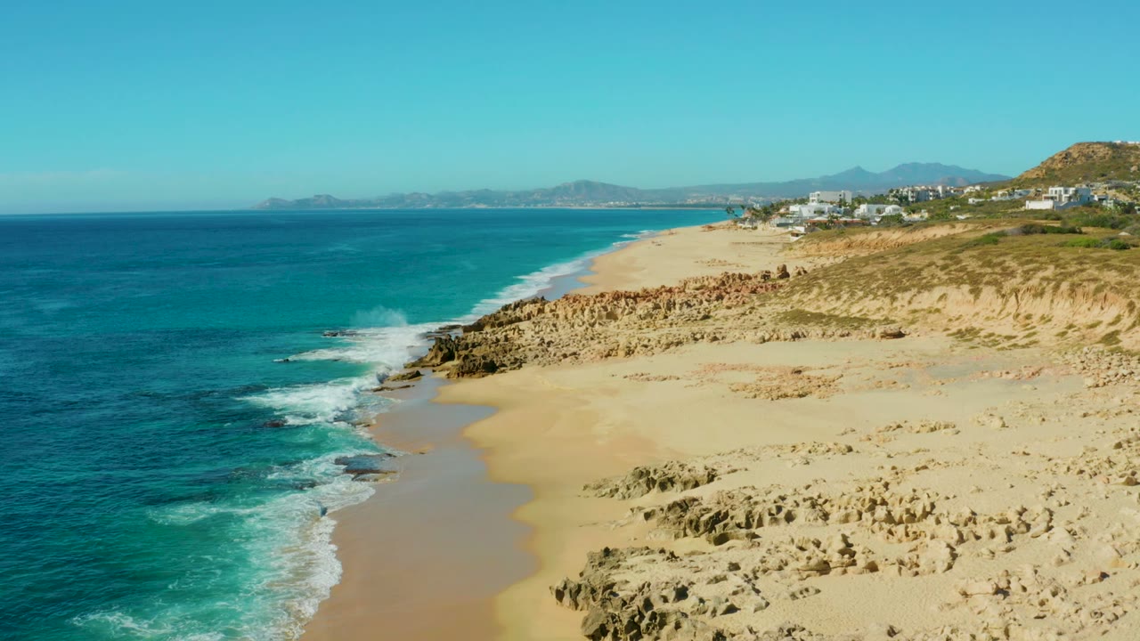 Opening aerial frame of Buzzards Beach East Cape in Baja California Sur, with turquoise water on the left, sand and rocks on the right, distant mountains, and no visible people.