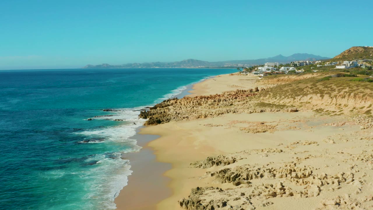 Buzzards Beach East Cape aerial view with a wider band of white surf near the lower left, tan sand and eroded rock formations across the right side, distant mountains, and no visible people.