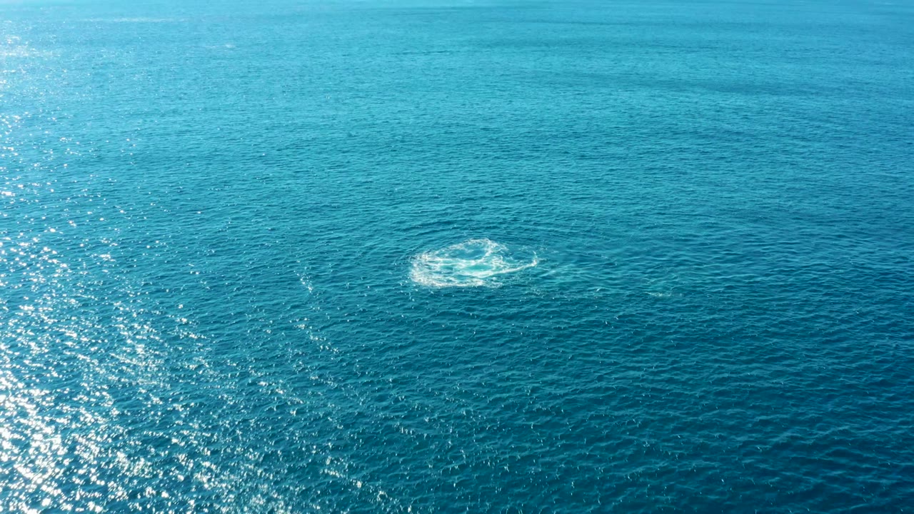 Small white splash ring on open blue ocean with vertical sunlight reflection.