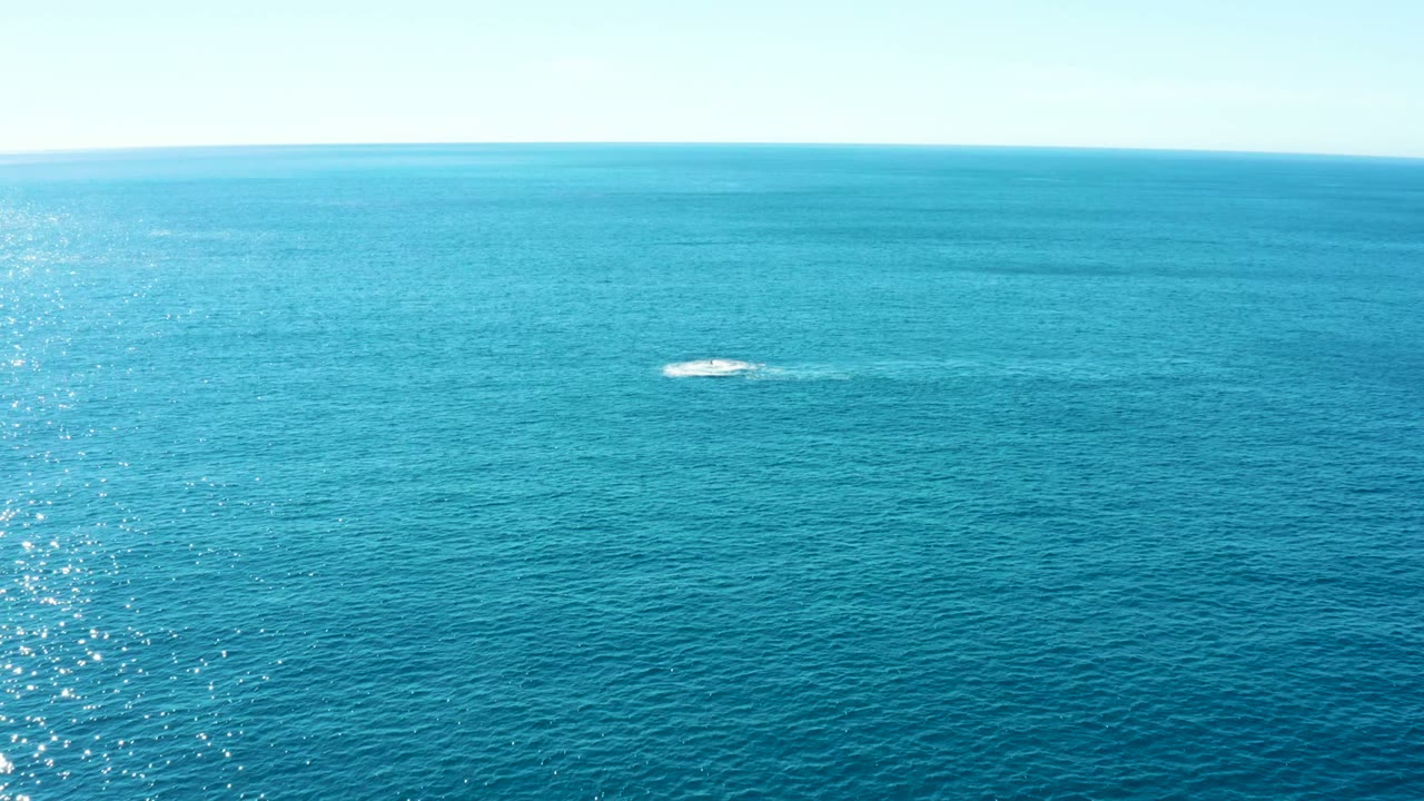 A wide aerial frame over Buzzards East Cape, Baja California Sur shows turquoise ocean, a small white whale spout near center, bright glare along the left water, and no visible people.