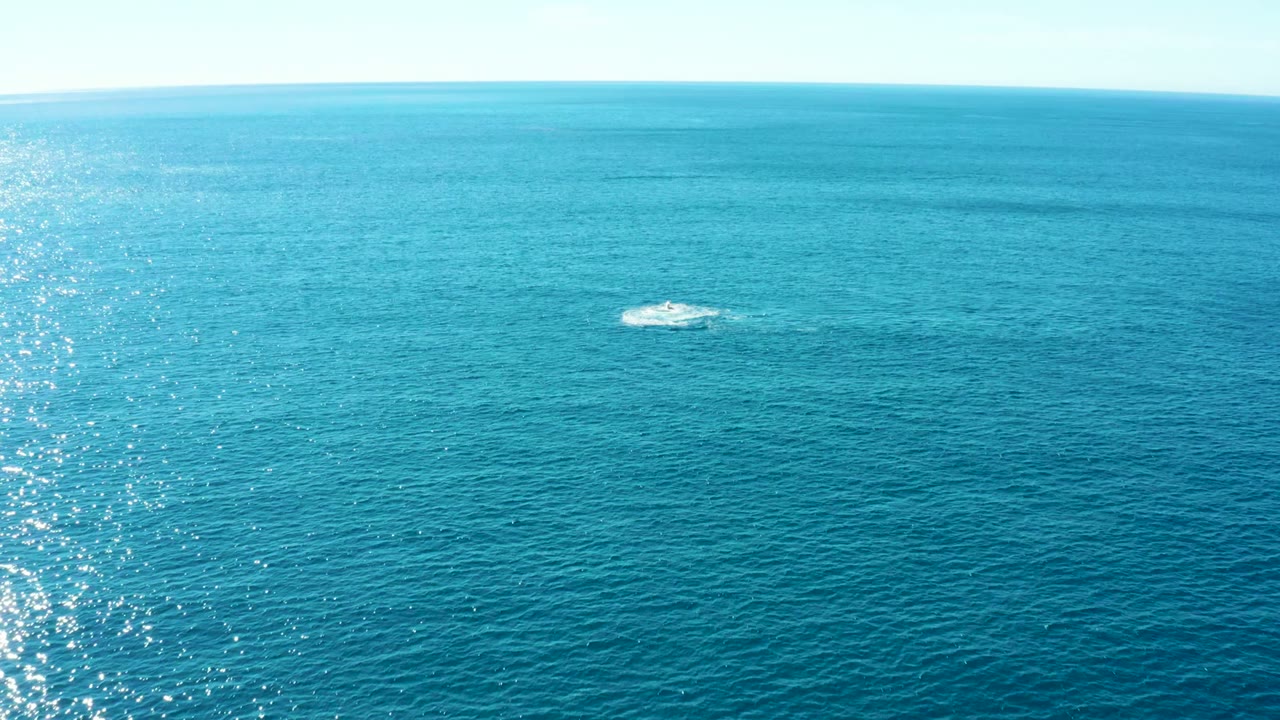 At 0:02, the frame shows Buzzards East Cape open ocean with a small white spout in the upper middle distance, textured teal water, bright reflection on the left, and no visible people.