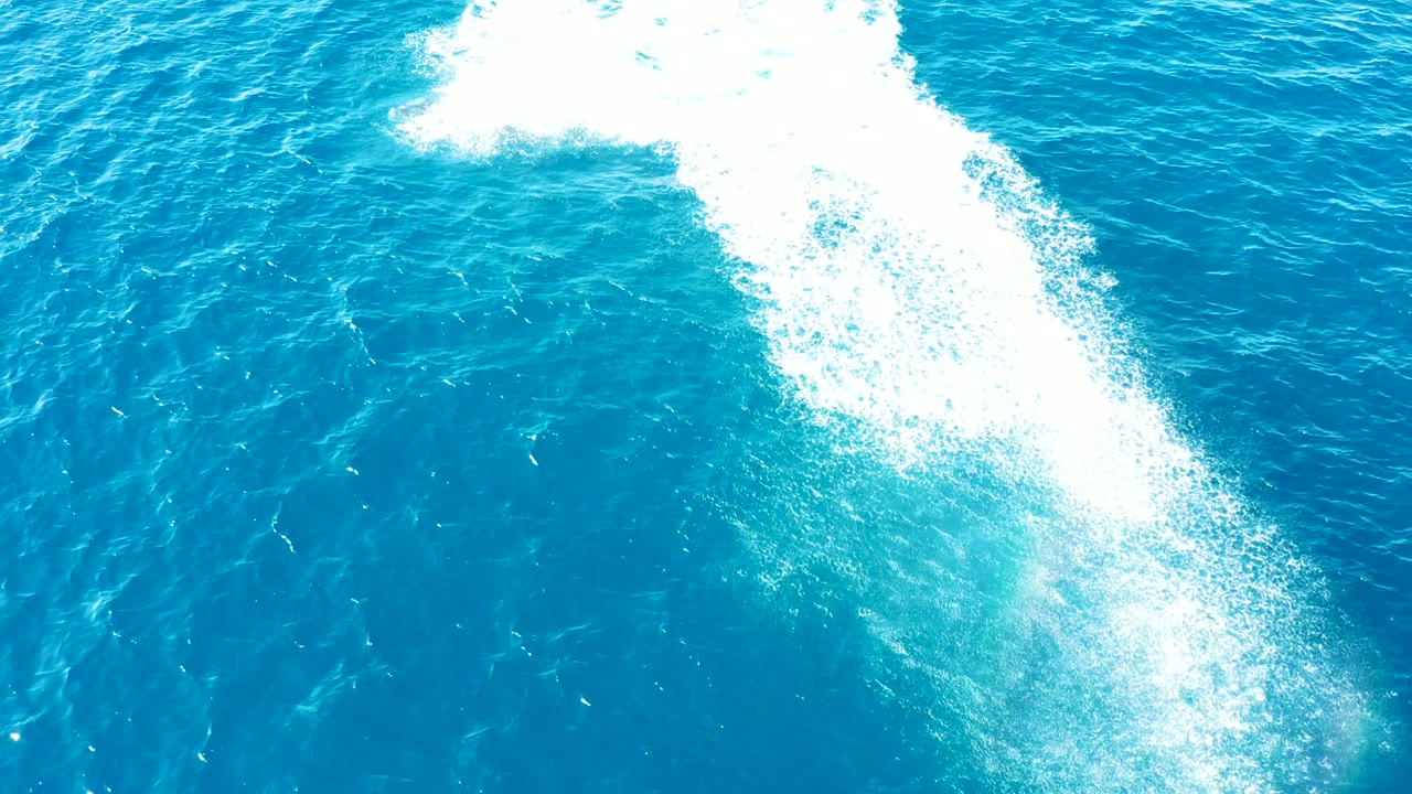 High overhead view in Baja California Sur with a stretched white wake-like spray pattern crossing bright blue water and no visible people.