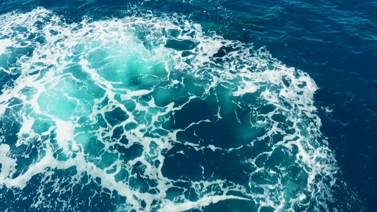 White foam curls around a bright turquoise patch in the open ocean near East Cape Los Cabos, with only faint underwater shape visible and no visible people.