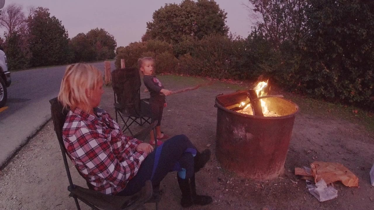 At Trestles Beach California, a blonde woman in a plaid shirt sits in the left foreground while a child sits near a burning metal fire pit on pavement.