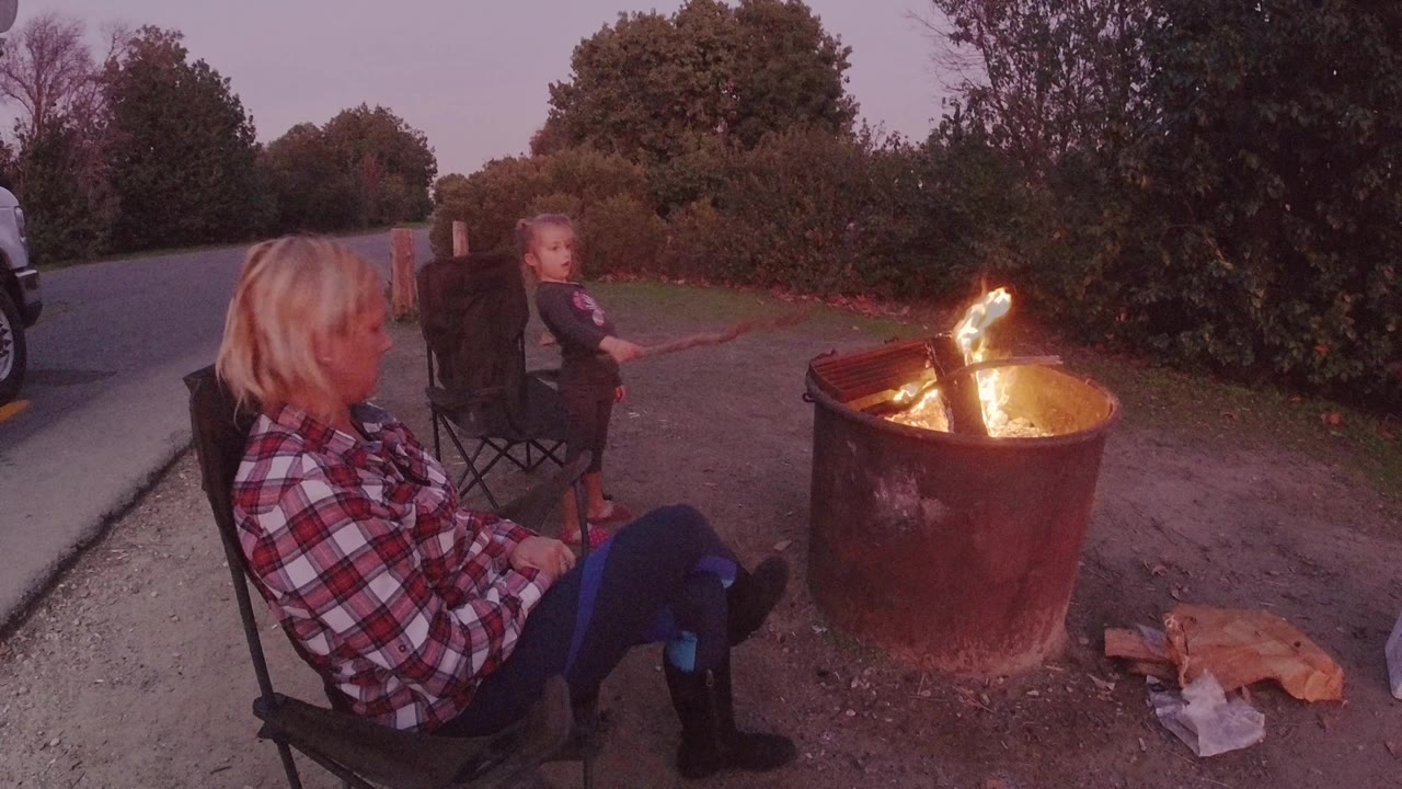 Woman in plaid shirt watches a fire pit as a child sits behind her.