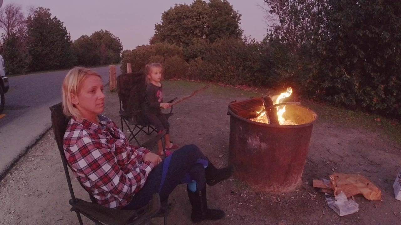 The woman looks toward the camera from a folding chair, with the child seated behind her and bright flames leaning over the rim of the fire pit.