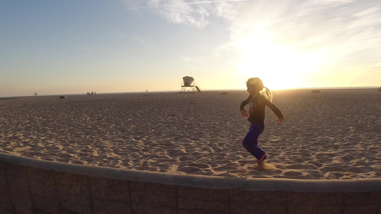 At Bolsa Chica California, a child in purple pants runs beside a low stone wall on a wide sandy beach while the sun flares near the horizon and a lifeguard tower sits in the distance.