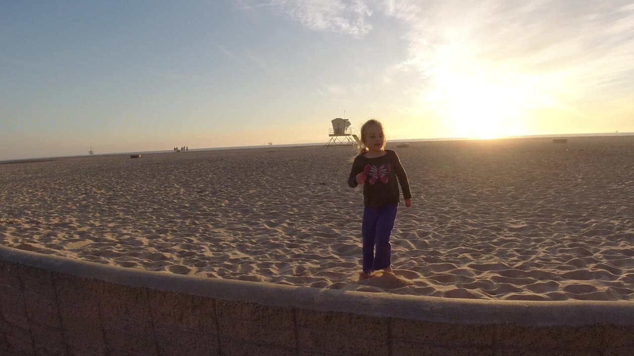 At Bolsa Chica California, a child stands nearer the camera by the low stone wall, partially silhouetted by the bright sunset over the sandy beach and distant lifeguard tower.