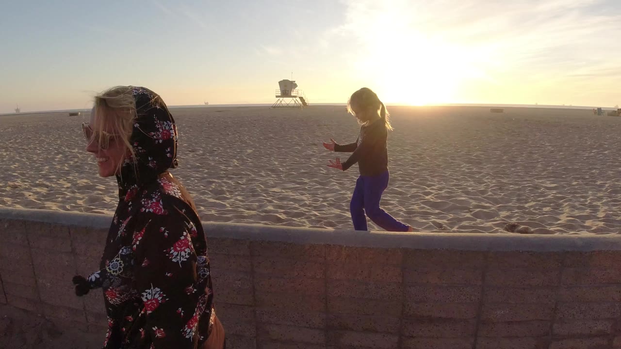 At Bolsa Chica California, a child walks along a low brick beach wall while an adult is partly visible close to camera and a lifeguard tower sits in the distance.