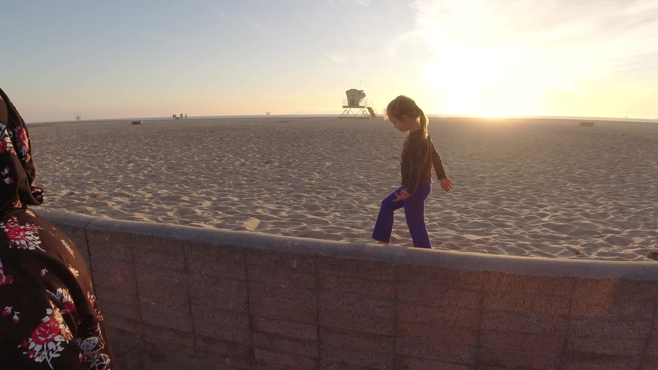 At Bolsa Chica California, the child remains on the wall while a close adult figure crosses the foreground edge, with the beach and lifeguard tower still visible beyond.