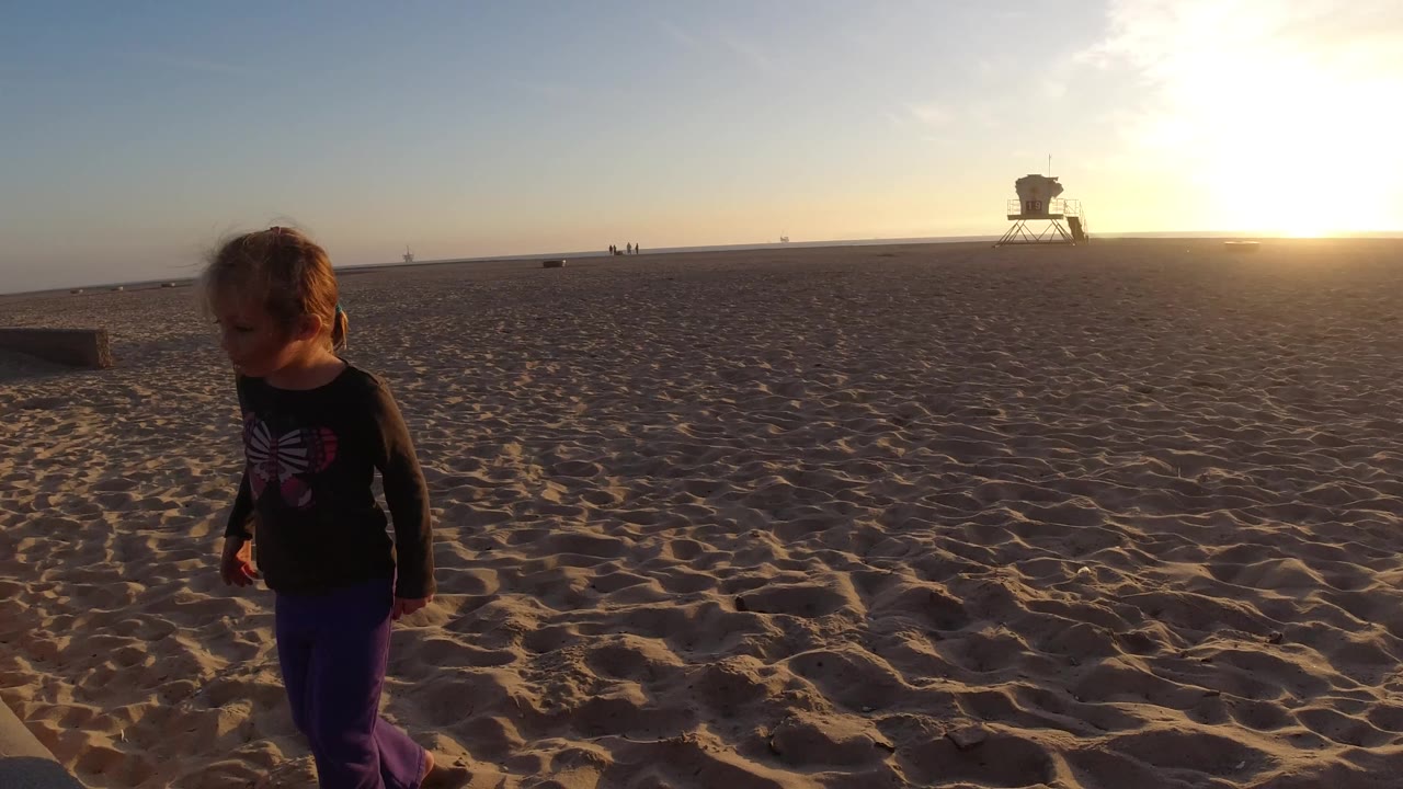 A blonde young girl stands near the left edge of a wide Bolsa Chica California beach frame, with rippled sand, a distant tower, and low sunset glare on the horizon.