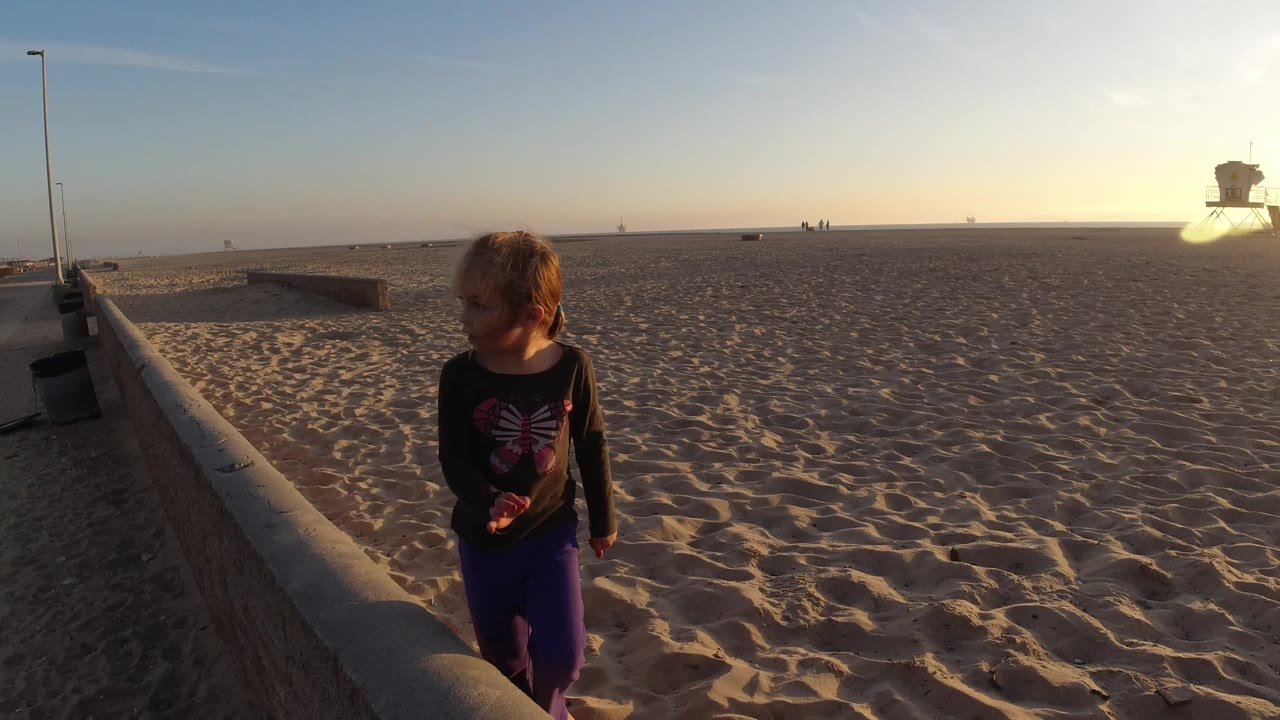A young girl stands near a concrete seawall at Bolsa Chica California, with the paved road at left, open sand at right, and a bright low sun across the horizon.