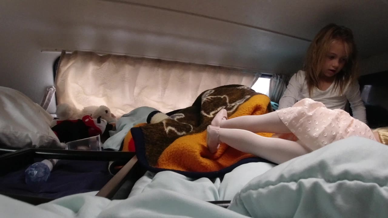 At 0:00, a young girl sits upright on a bed in Santa Maria California while a doll in a pale pink dress lies across blankets near beige curtains.