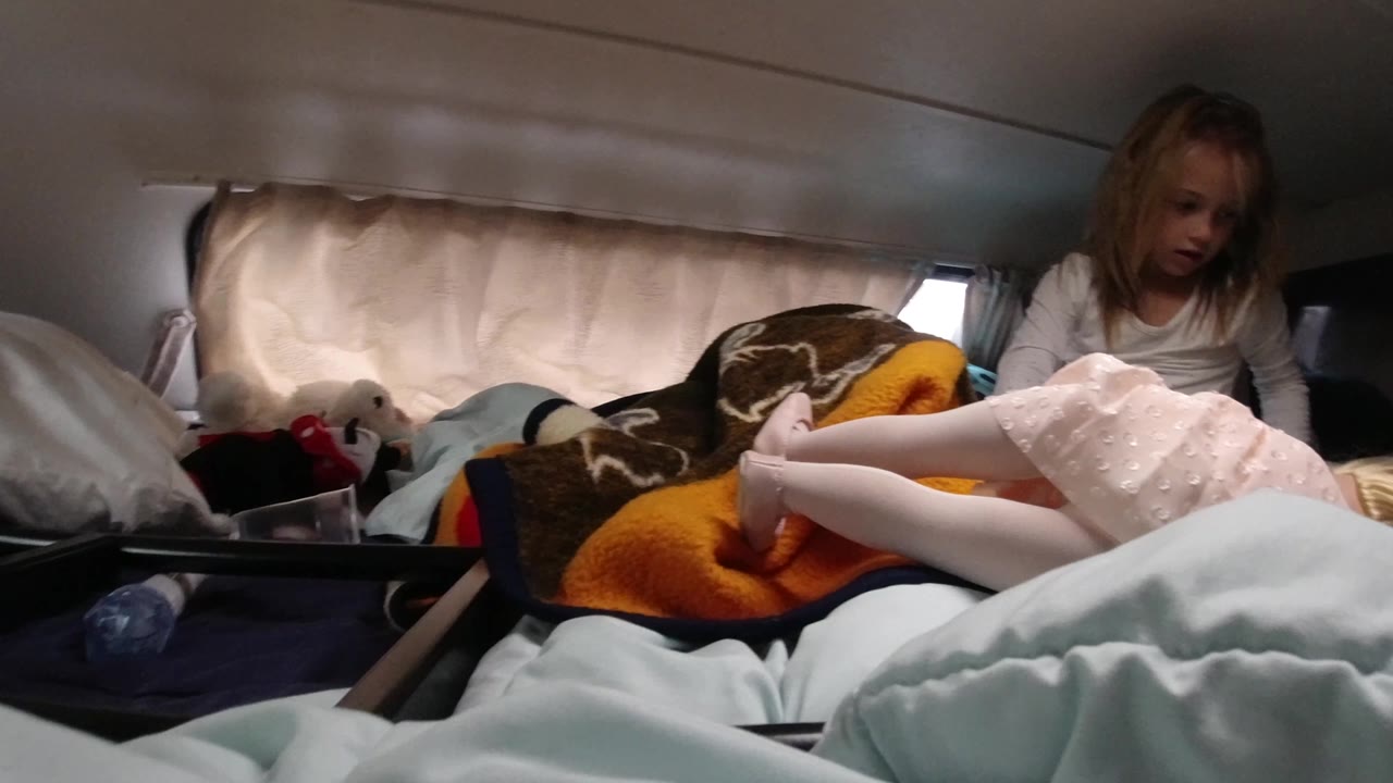 Seated child watches a doll lying on a bed with blankets and toys nearby.