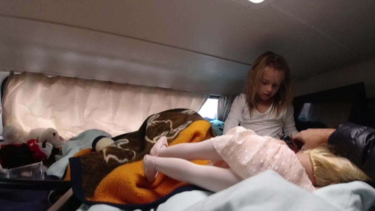 At 0:02, an adult hand enters from the right to adjust or hold a doll as the seated girl watches from the bed inside the Santa Maria California interior.