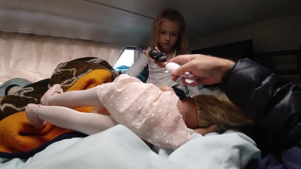 In Santa Maria California, an adult hand holding a small toy appears above the child in the pink dress as another child watches from the rear of the vehicle bed.