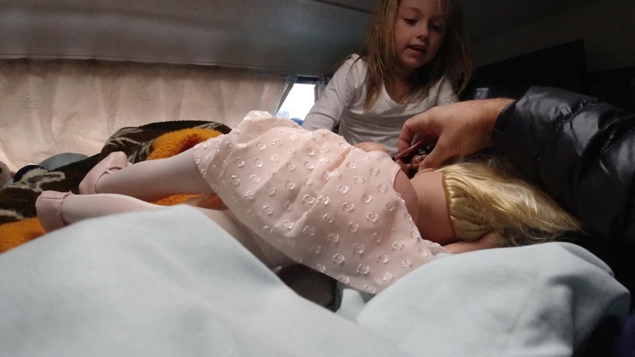 The seated girl is more visible above the bedding as an adult hand works near the child lying in the pink dress inside the dim Santa Maria California interior.