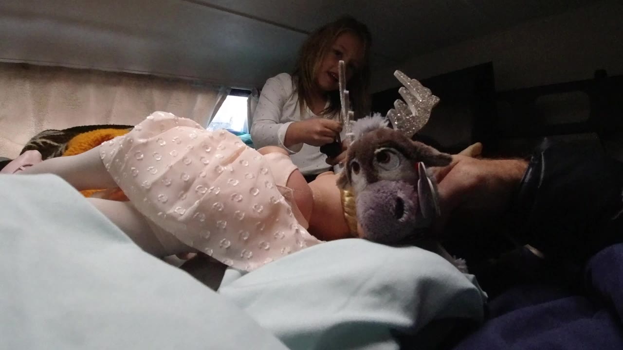 In Santa Maria California, a young child in a pink dress lies across bedding as an adult hand holds a plush reindeer toy near her face and a woman sits behind her.
