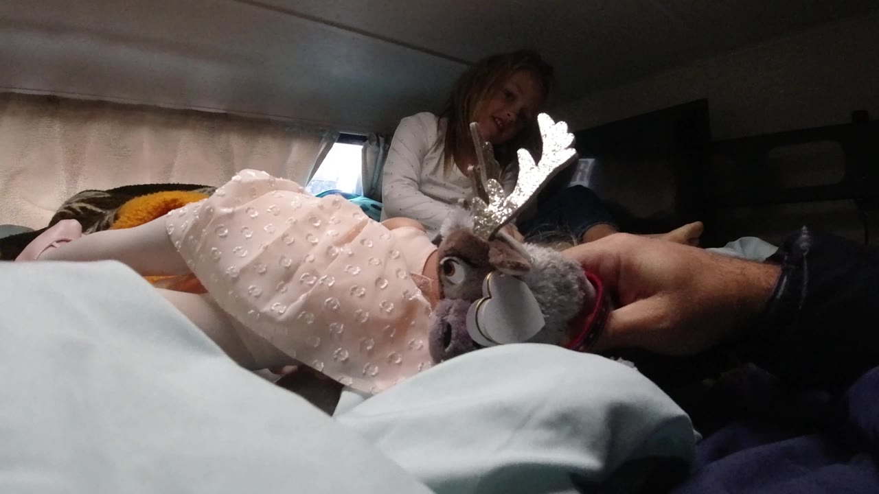 A hand raises the plush reindeer toy beside the child on the bed, with the woman slightly farther back and a small window brightening the dim camper interior.