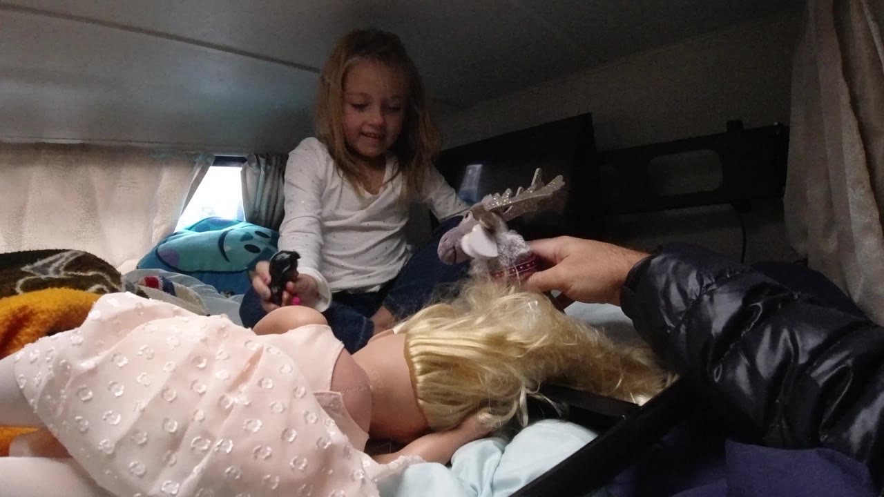 In Santa Maria California, the girl smiles while holding a small dark toy, with the blonde doll lying close to camera and the reindeer toy lifted by an adult hand.