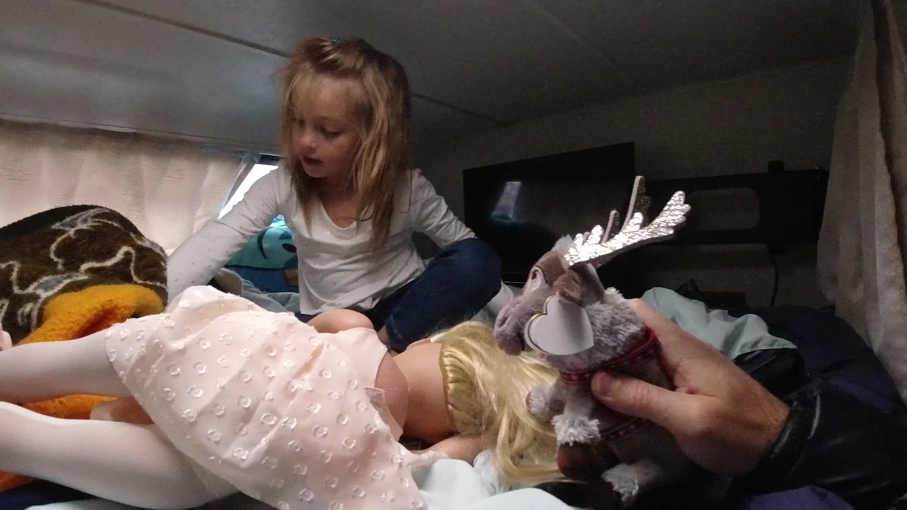 Girl leaning over a bed while a plush reindeer is held near a doll.